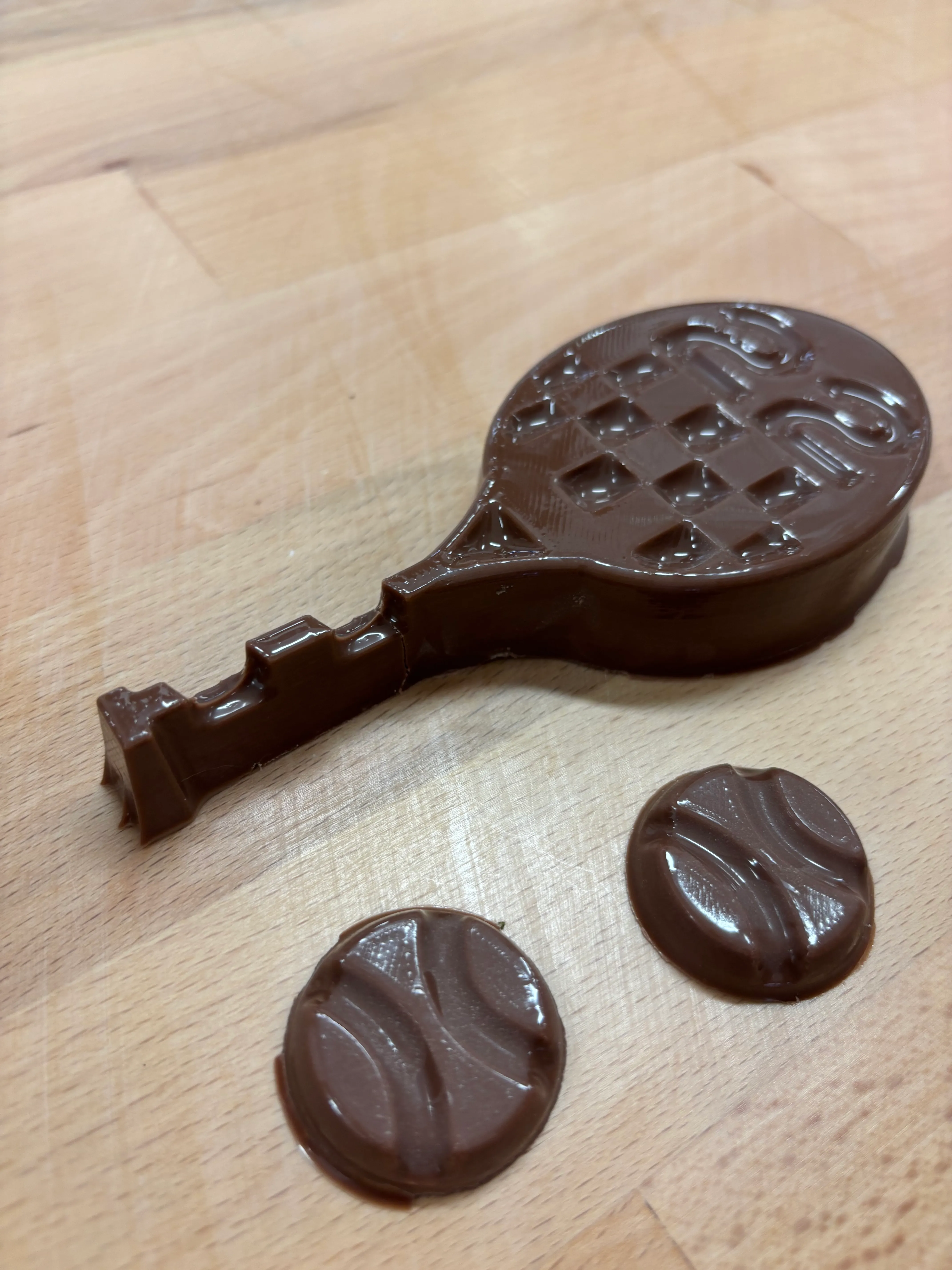 Three finished chocolate pieces — a racquet with a raised 22 and two ball coins — laid out on the work surface.