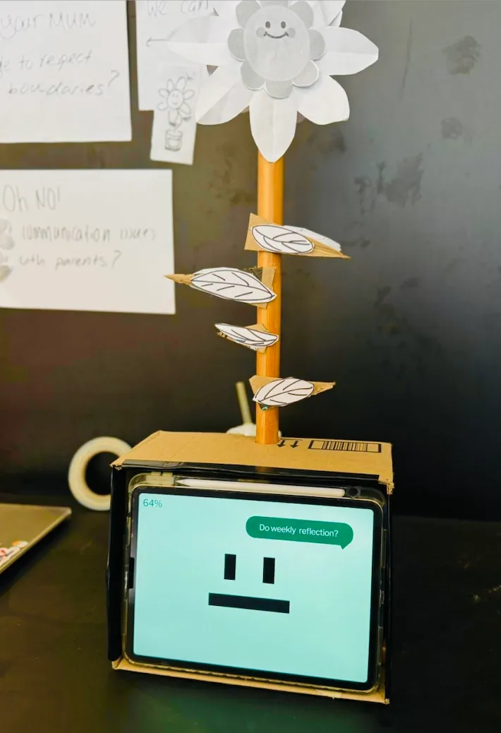 The first Mira prototype — a paper flower on a taped pole rising from a small cardboard planter, with a tablet mounted at the base showing a simple face.